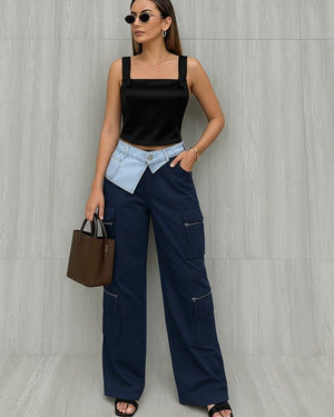 Woman wearing a black top, navy pants, and sunglasses against a gray wall.
