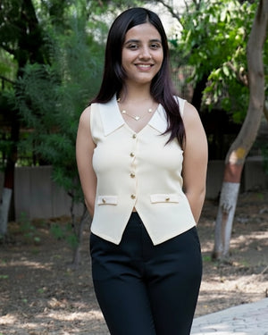 Woman wearing a cream vest over a black top outdoors with greenery in the background