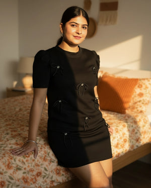 Woman in a black dress standing in a bedroom with floral bedding and a lamp.