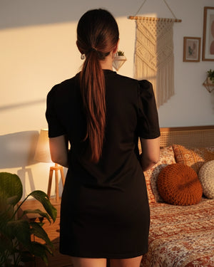 Woman in a black dress standing in a cozy room with warm lighting.
