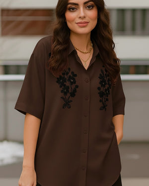 Woman wearing a brown blouse with black floral embroidery against a blurred background