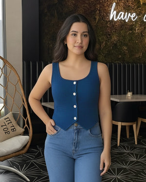 Woman wearing a blue sleeveless top with white buttons and jeans in an indoor setting.