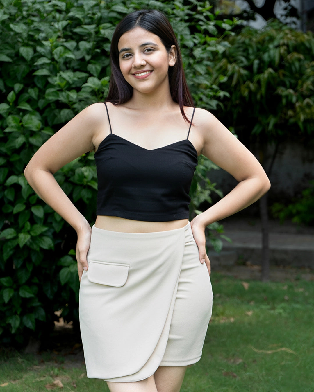 Woman wearing a black top and beige skirt standing outdoors with greenery in the background