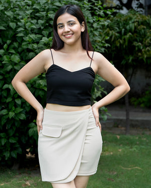 Woman wearing a black top and beige skirt standing outdoors with greenery in the background