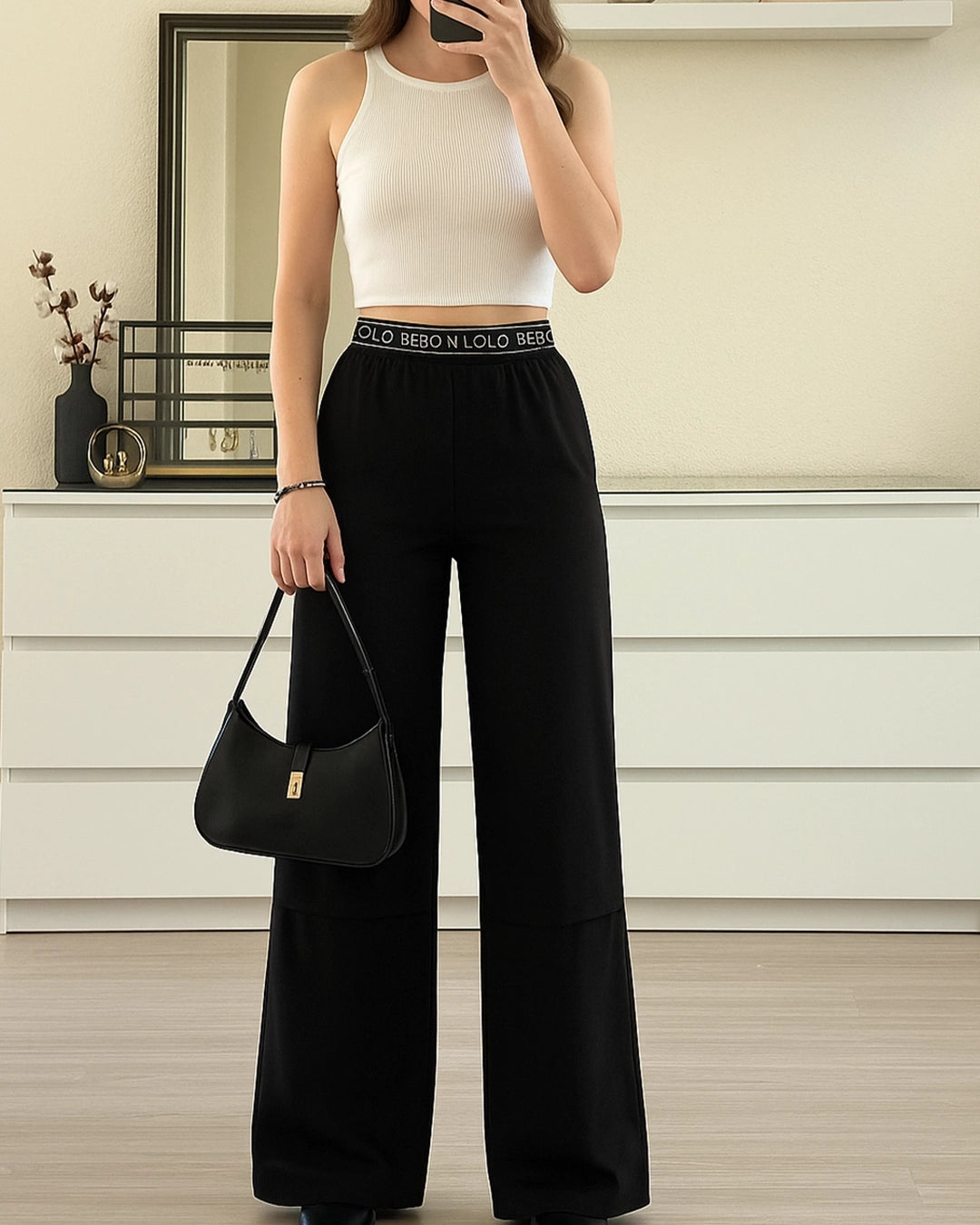 Person wearing a white tank top and black wide-leg pants with a belt, holding a black handbag.