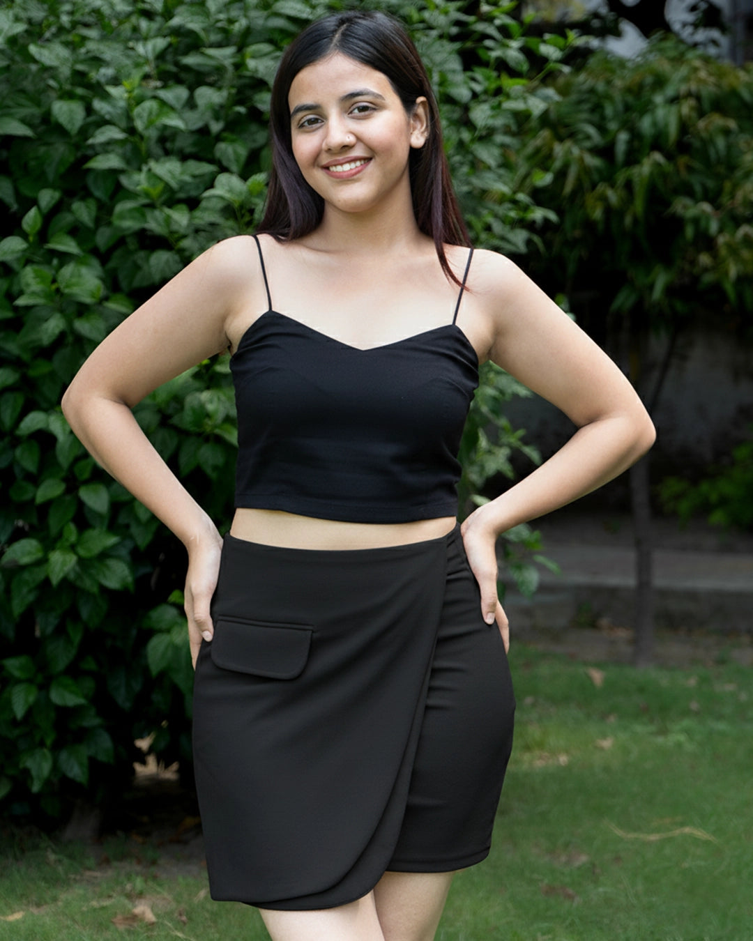 Woman wearing a black top and skirt standing outdoors with greenery in the background