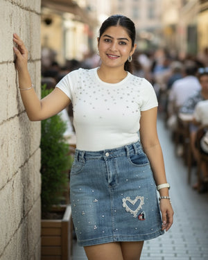 Woman in a white top and blue denim skirt standing against a wall in an outdoor setting.