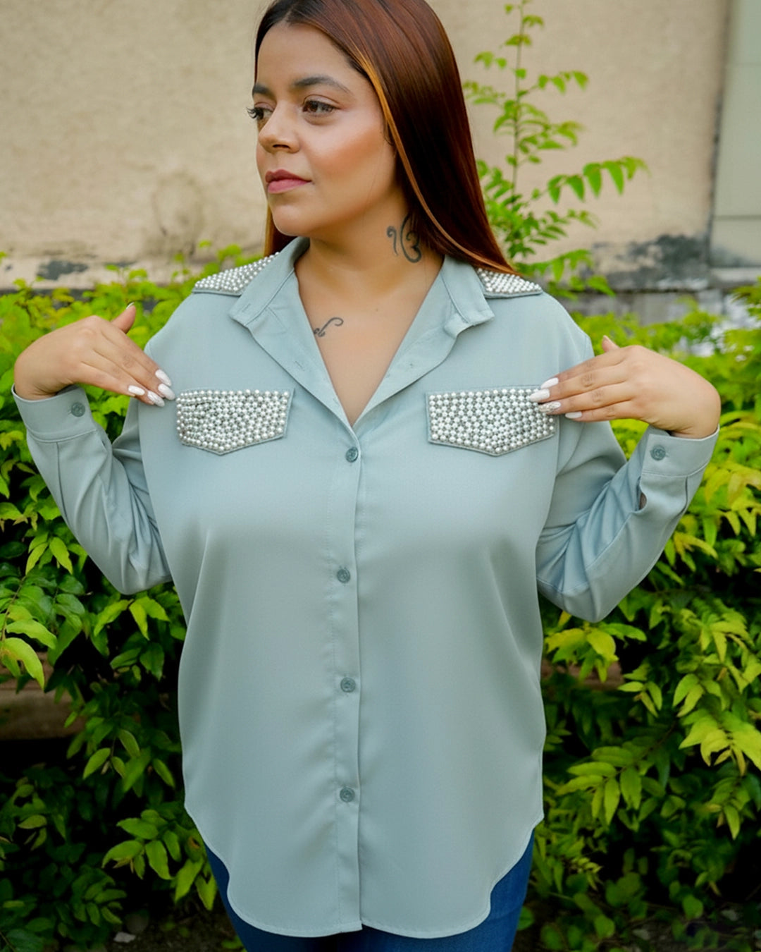 Woman wearing a light blue shirt with embellished shoulder pads in front of green foliage.