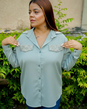 Woman wearing a light blue shirt with embellished shoulder pads in front of green foliage.