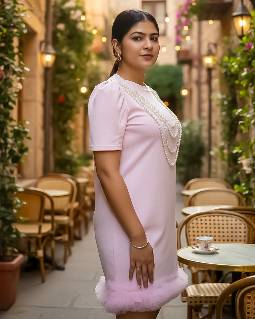Woman in a light pink dress with white embellishments standing in an outdoor cafe setting.
