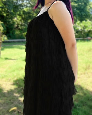 Person wearing a black fringe vest outdoors with greenery in the background