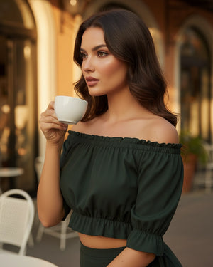 Woman in a green off-shoulder top holding a white mug outdoors.