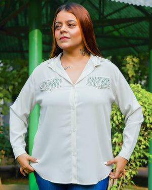 Woman wearing a light gray shirt with decorative elements, standing outdoors with greenery in the background.