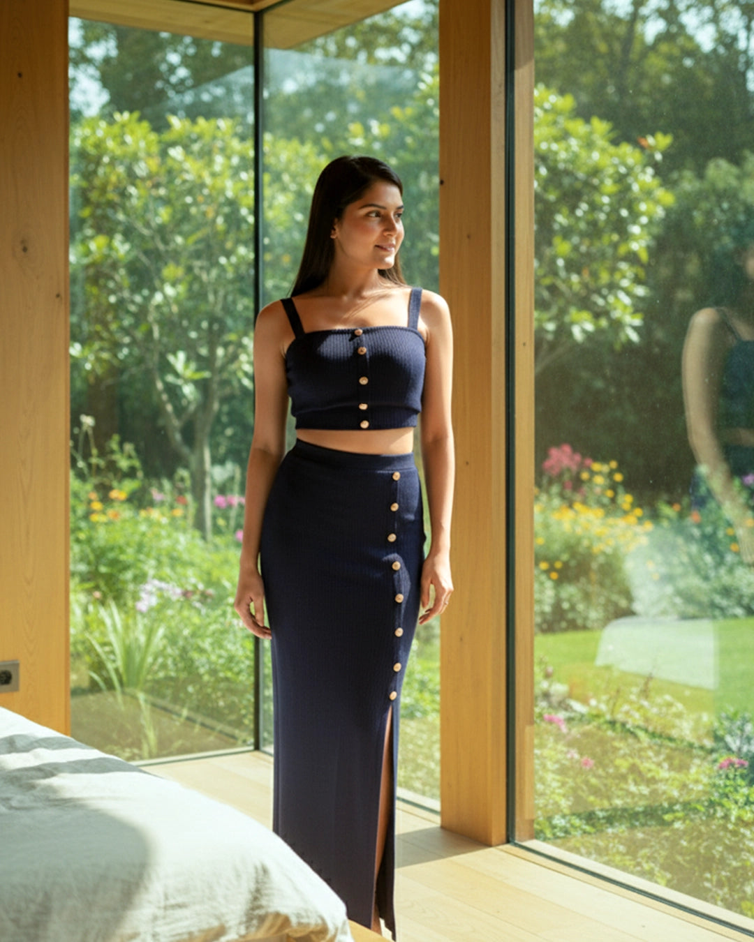 Woman in a navy blue outfit standing in a room with large windows showing a garden view.