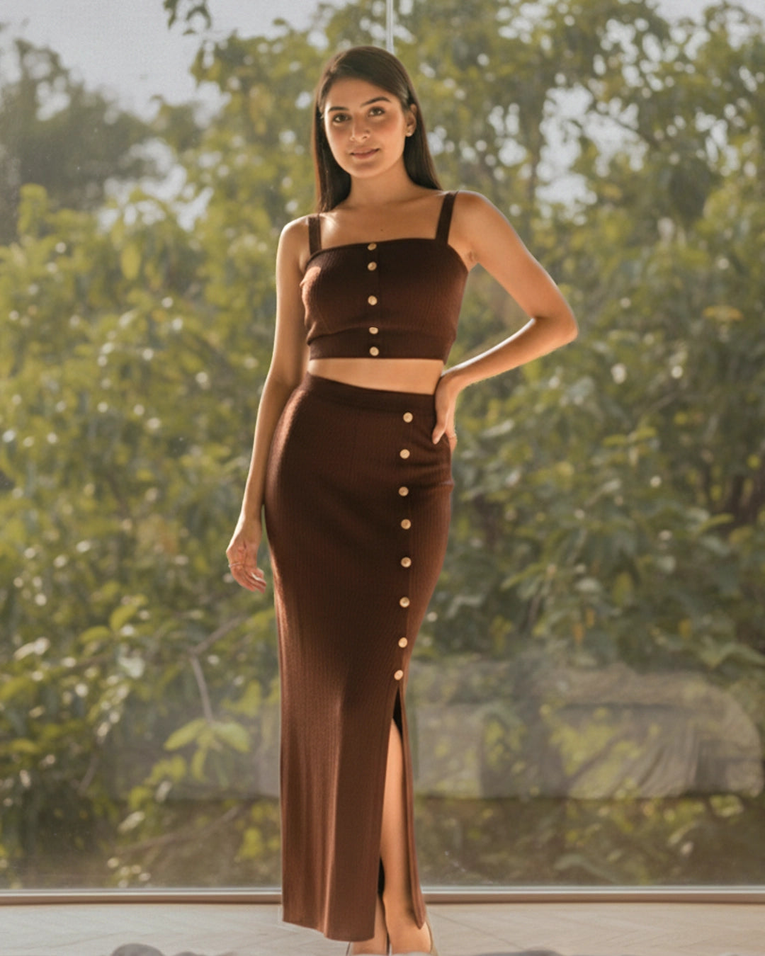Woman wearing a brown outfit with button details against a blurred green outdoor background