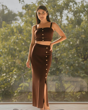 Woman wearing a brown outfit with button details against a blurred green outdoor background