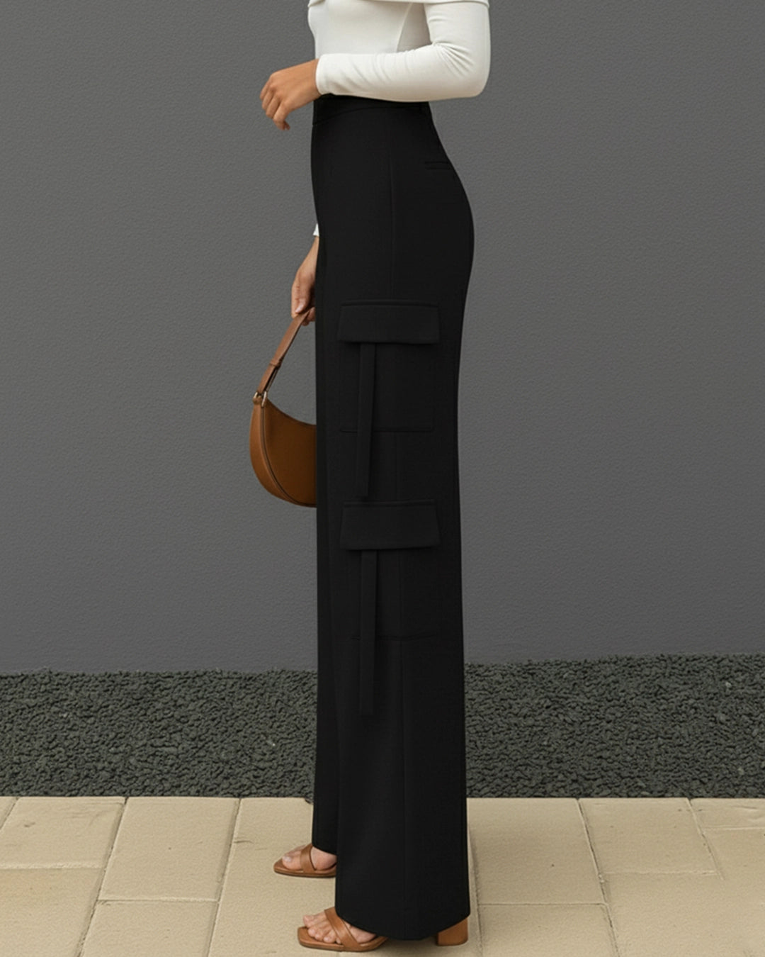 Person wearing black cargo pants and a white top, holding a brown handbag against a gray background.