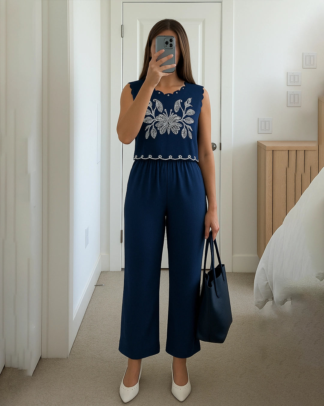 Person wearing a navy blue jumpsuit with white floral embroidery, holding a black handbag indoors.