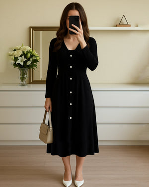 Woman wearing a black dress with white buttons, holding a beige handbag indoors.