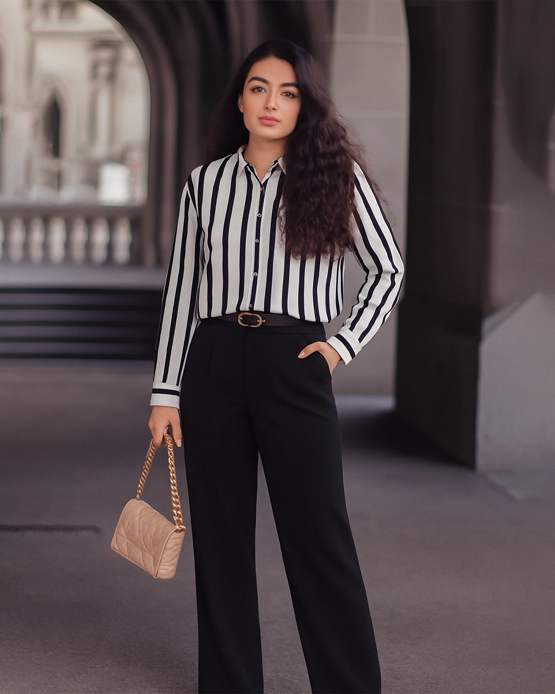 Woman wearing a black and white striped shirt with black pants, holding a beige handbag in an outdoor setting.