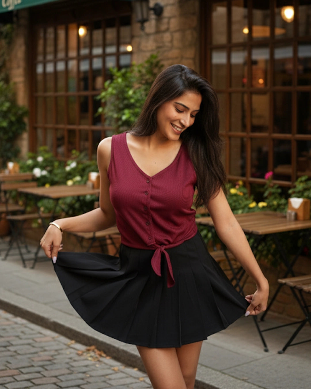 Woman in a red top and black skirt standing outdoors in front of a building with tables and chairs.
