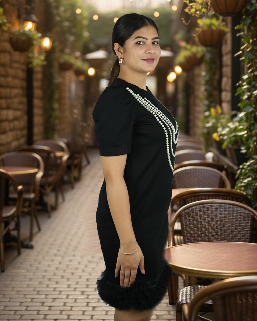 Woman in a black dress with white embellishments standing in an outdoor setting with tables and chairs.