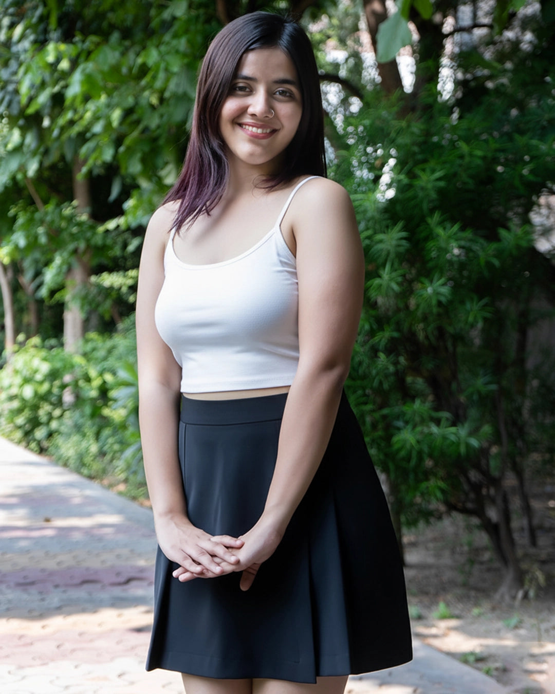 Woman wearing a white tank top and black skirt standing outdoors with greenery in the background