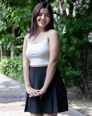 Woman wearing a white tank top and black skirt standing outdoors with greenery in the background