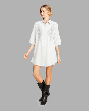 Woman wearing a white dress with floral embroidery and black boots on a gray background