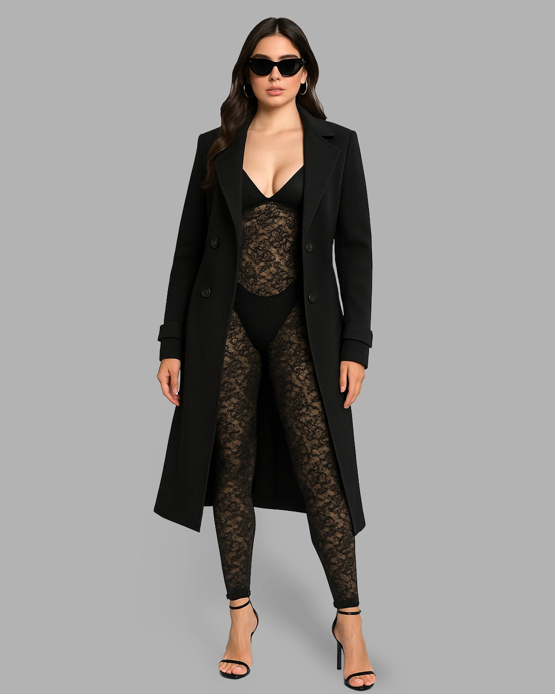 Woman wearing a long black coat over a sheer black top and leggings on a gray background