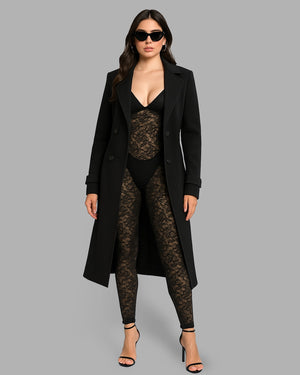Woman wearing a long black coat over a sheer black top and leggings on a gray background