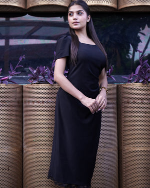 Woman in a black dress standing against a textured wall with plants.