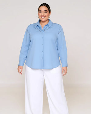 Woman wearing a light blue button-up shirt with decorative studs and white pants on a plain background